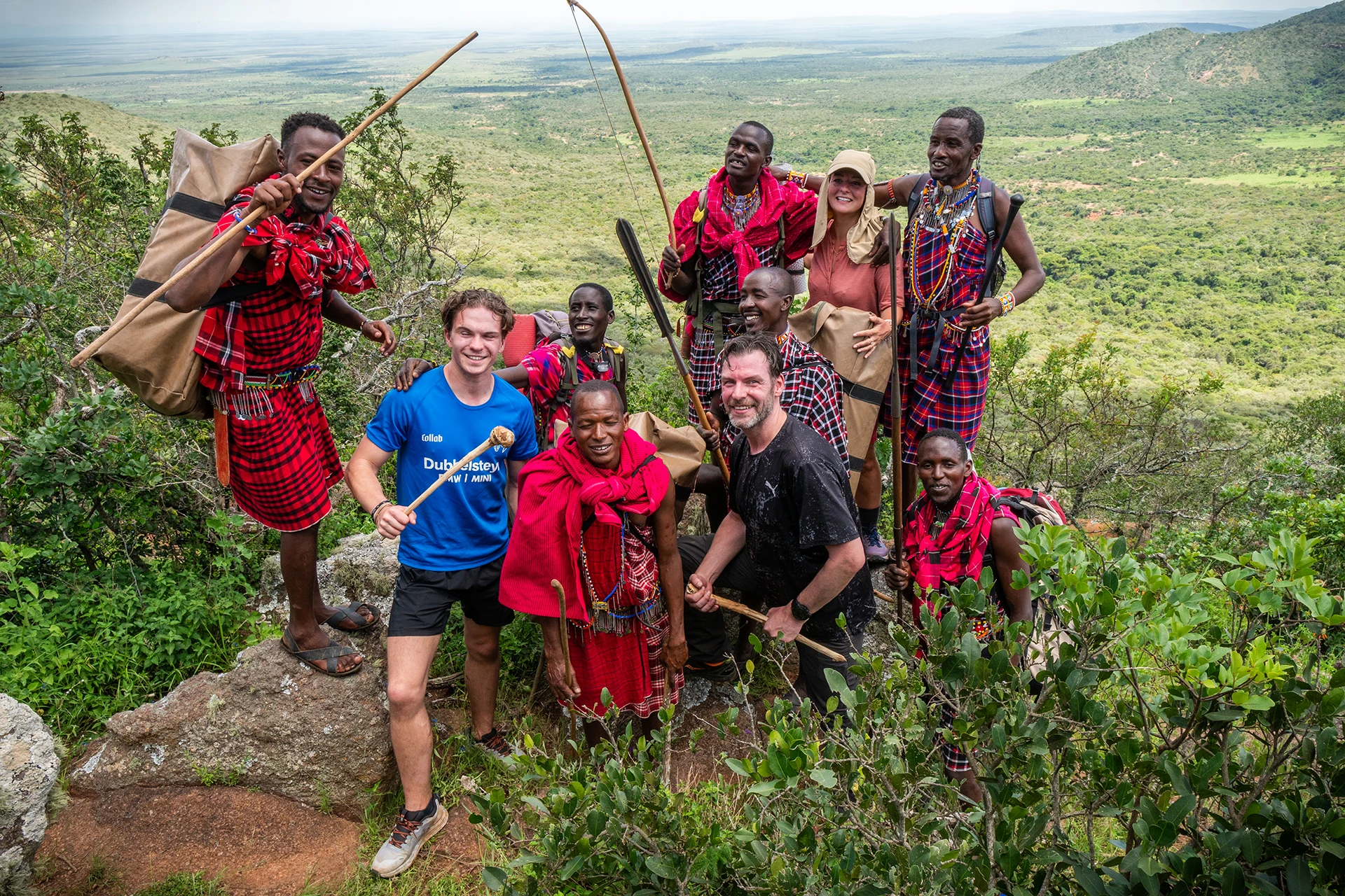 Groep mensen in traditionele kleding poseert samen met twee wandelaars in groene heuvels