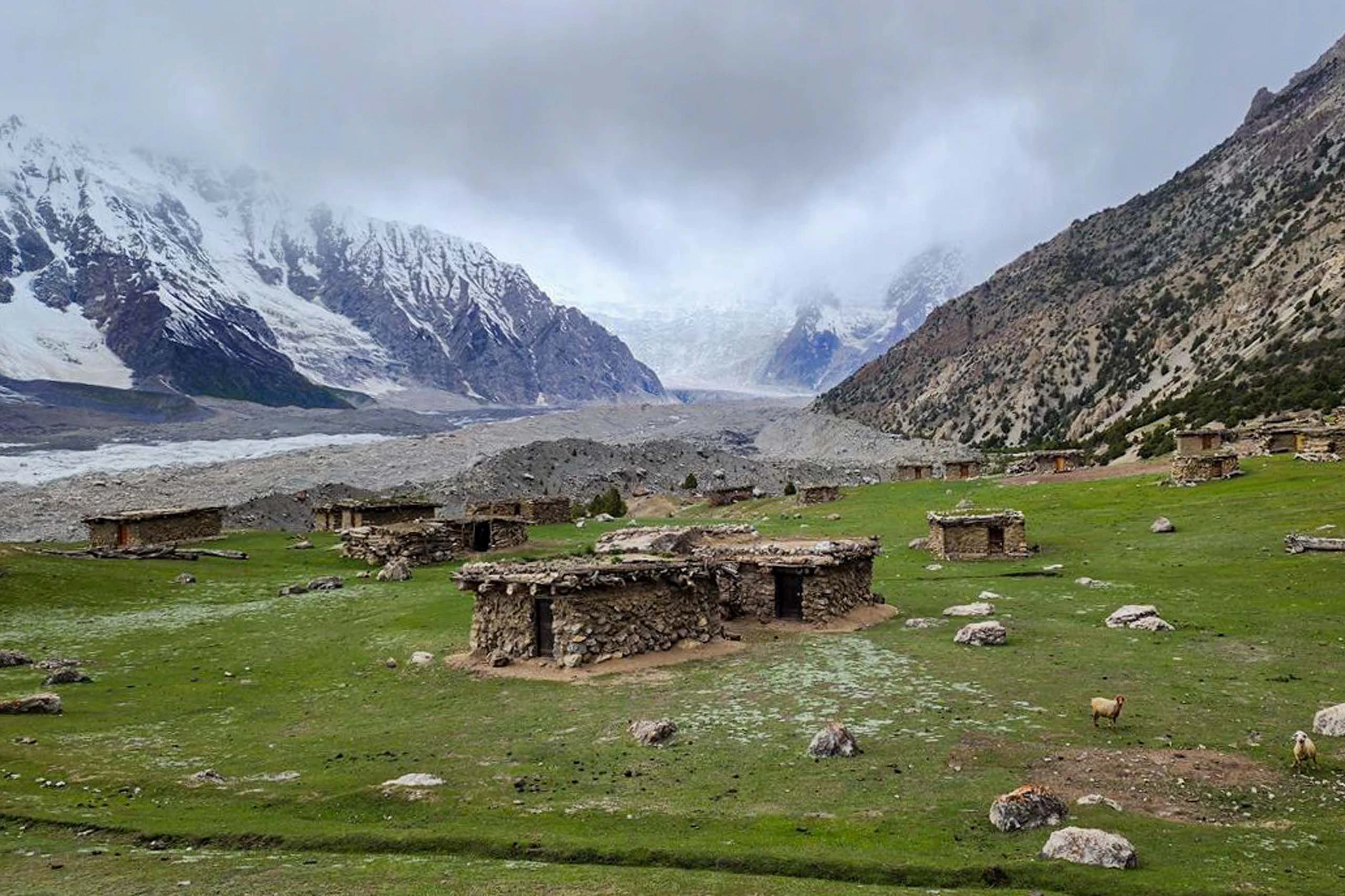 Bergweide met stenen huisjes en besneeuwde toppen onder bewolkte lucht