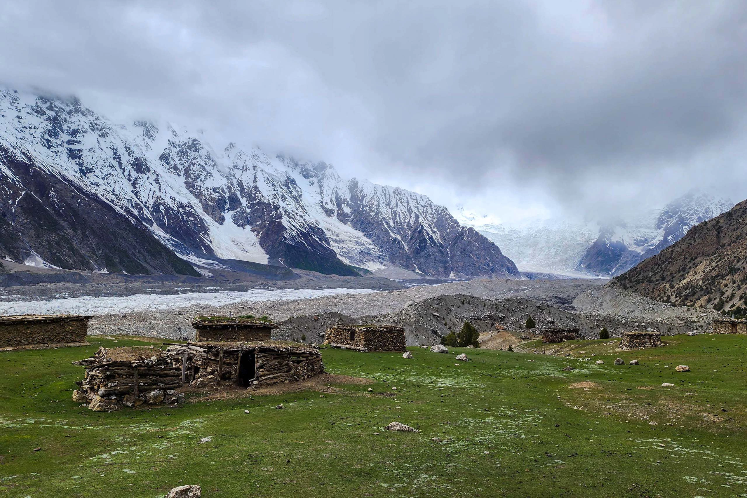 Groene alpenweide met stenen hut en besneeuwde bergtoppen onder bewolkte lucht
